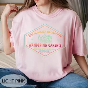May include: Light pink t-shirt with a colorful graphic that reads "BIG SUMMER BLOWOUT, WANDERING OAKEN'S, Trading Post & Sauna, ARENDELLE". The design features a cabin and mountain scene within a hexagon shape.