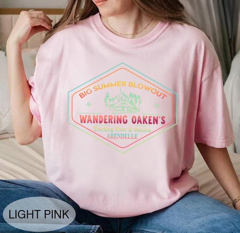 May include: Light pink t-shirt with a colorful graphic that reads "BIG SUMMER BLOWOUT, WANDERING OAKEN'S, Trading Post & Sauna, ARENDELLE". The design features a cabin and mountain scene within a hexagon shape.