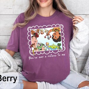 K&ouml;nnte beinhalten: Beerenfarbenes T-Shirt mit einem gerahmten Grafikmotiv von Toy Story-Charakteren, darunter Woody und Buzz Lightyear. Der Text "You've got a friend in me" steht darunter. Das T-Shirt hat einen Rundhalsausschnitt.