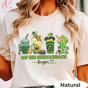Puede incluir: Una camiseta de color natural con un diseño del Día de San Patricio. El gráfico incluye personajes de dibujos animados, bebidas verdes y la frase "Let the Shenanigans Begin" en texto verde con un corazón.
