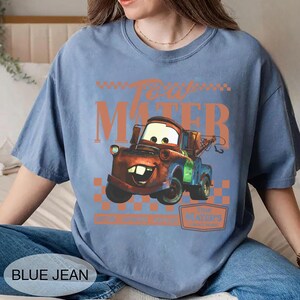 Puede incluir: Camiseta azul claro con un dise&ntilde;o de Mate de la pel&iacute;cula Cars. El dise&ntilde;o incluye el texto "Tow Mater" en naranja, con una imagen de dibujos animados del personaje. La camiseta tambi&eacute;n tiene el texto "Blue Jean" en la parte inferior.