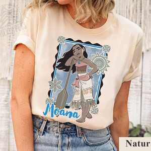 May include: Cream-coloured t-shirt featuring a cartoon illustration of Moana with a paddle. The design includes a blue frame, floral accents, and the name "Moana" in blue script. The word "Natural" is printed at the bottom.