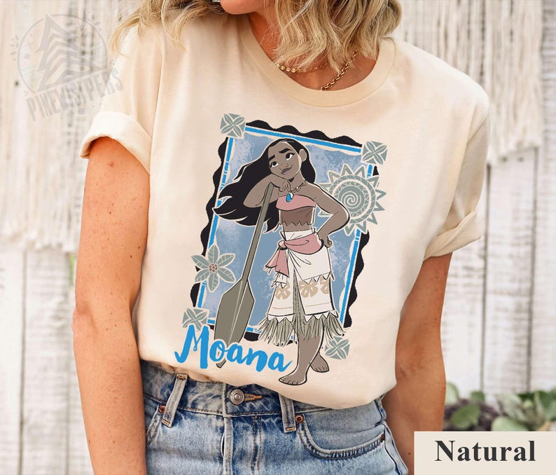 May include: Cream-coloured t-shirt featuring a cartoon illustration of Moana with a paddle. The design includes a blue frame, floral accents, and the name "Moana" in blue script. The word "Natural" is printed at the bottom.