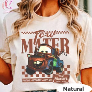 Puede incluir: Una camiseta de color natural con un gr&aacute;fico de Mate de la pel&iacute;cula Cars. El dise&ntilde;o incluye el texto "Tow Mater" en marr&oacute;n, con la imagen de Mate y la frase "Anytime - Anywhere - Anyplace".