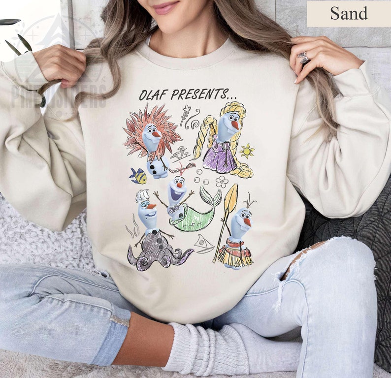Puede incluir: Sudadera de color arena con un gr&aacute;fico colorido de Olaf de Frozen con varios disfraces, con el texto "OLAF PRESENTS...". El dise&ntilde;o incluye a Olaf como sirena, pirata y otros personajes. La sudadera es de estilo casual y c&oacute;modo.