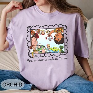 K&ouml;nnte beinhalten: Orchideenfarbenes T-Shirt mit einem gerahmten Cartoon-Bild von Charakteren aus dem Film Toy Story. Der Rahmen hat einen gewellten Rand, und der Text "You've got a friend in me" steht darunter. Das Wort "ORCHID" ist unten aufgedruckt.