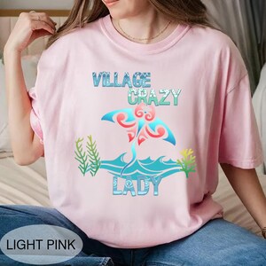 K&ouml;nnte beinhalten: Hellrosa T-Shirt mit dem Schriftzug "VILLAGE CRAZY LADY" in Blau und Gr&uuml;n. Das Design zeigt einen Mantarochen mit blauen und roten Akzenten, vor einem wellenf&ouml;rmigen Ozean-Design. Unten steht "LIGHT PINK".