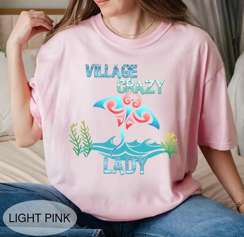 K&ouml;nnte beinhalten: Hellrosa T-Shirt mit dem Schriftzug "VILLAGE CRAZY LADY" in Blau und Gr&uuml;n. Das Design zeigt einen Mantarochen mit blauen und roten Akzenten, vor einem wellenf&ouml;rmigen Ozean-Design. Unten steht "LIGHT PINK".