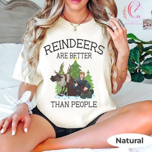 May include: A cream-colored t-shirt with the text "REINDEERS ARE BETTER THAN PEOPLE" in black. The graphic features a cartoon reindeer, a man playing a lute, and Christmas trees. The shirt is labeled "Natural" at the bottom.