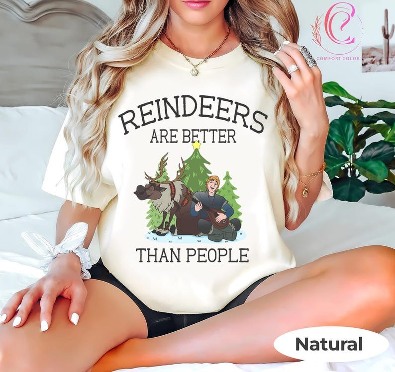May include: A cream-colored t-shirt with the text "REINDEERS ARE BETTER THAN PEOPLE" in black. The graphic features a cartoon reindeer, a man playing a lute, and Christmas trees. The shirt is labeled "Natural" at the bottom.