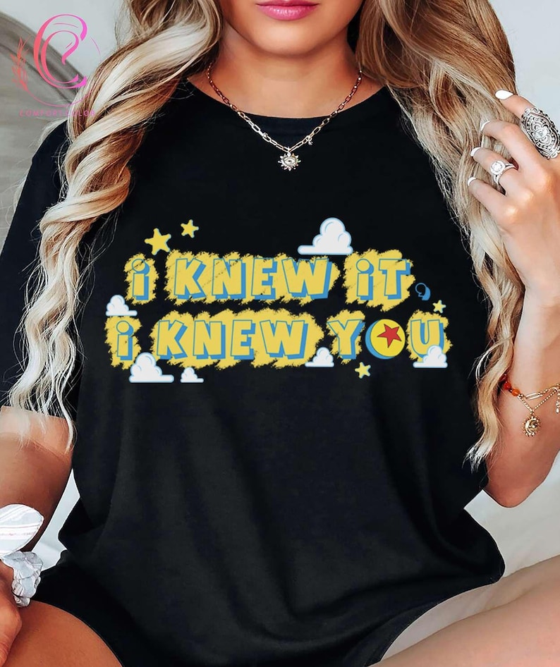 May include: A black short-sleeved t-shirt featuring the text "I KNEW IT I KNEW YOU" in blue letters on a yellow textured background. The design includes small white clouds and yellow stars, with a red star inside the letter O in YOU.