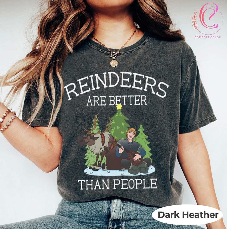 May include: Dark heather gray t-shirt with the text "REINDEERS ARE BETTER THAN PEOPLE". The shirt features a graphic of a reindeer, a man playing a lute, and Christmas trees. The shirt is made of Comfort Colors material.