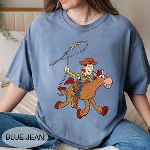 May include: A light blue t-shirt featuring a cartoon illustration of a cowboy riding a horse. The cowboy is wearing a hat and holding a lasso. The word "John" is written above the cowboy. The words "BLUE JEAN" are at the bottom.