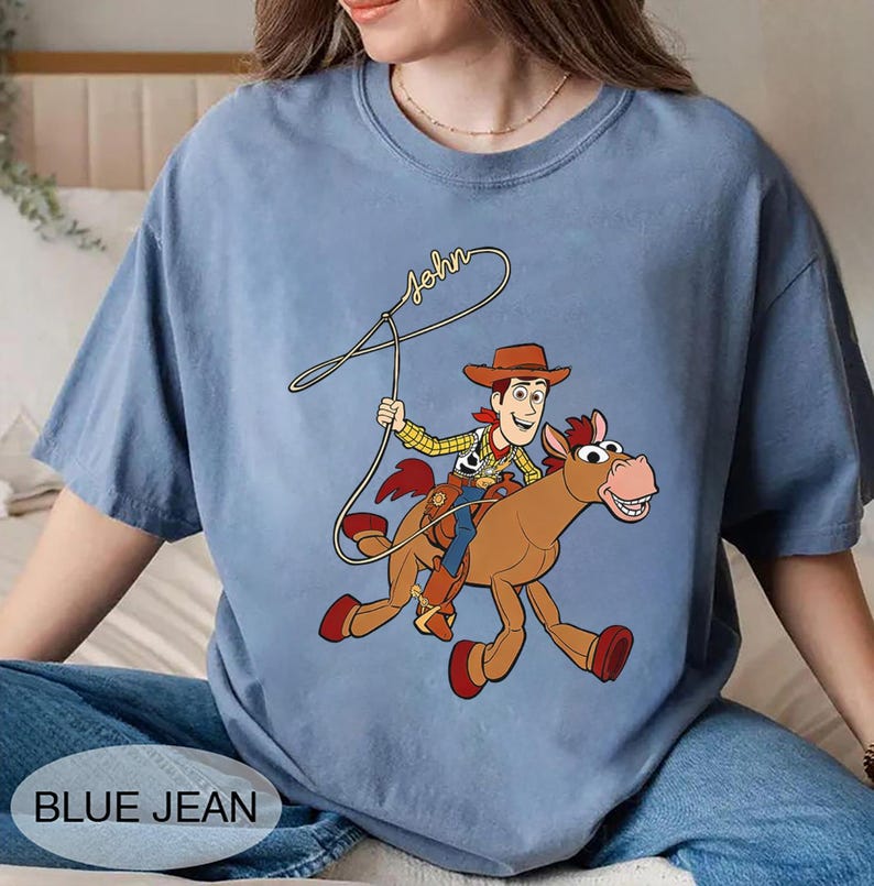 May include: A light blue t-shirt featuring a cartoon illustration of a cowboy riding a horse. The cowboy is wearing a hat and holding a lasso. The word "John" is written above the cowboy. The words "BLUE JEAN" are at the bottom.