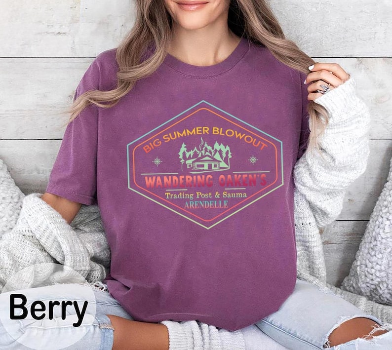 May include: Berry-colored t-shirt with a graphic that reads "Big Summer Blowout, Wandering Oaken's Trading Post & Sauma, Arendelle." The design is in a hexagon shape with a cabin and trees. The shirt is a crew neck.