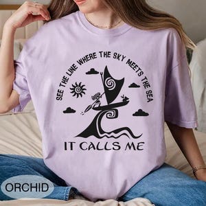 以下が含まれることがあります： オーキッド色のTシャツ。黒いグラフィックで、波に乗るヨット、太陽、雲、そして「SEE THE LINE WHERE THE SKY MEETS THE SEA IT CALLS ME」というテキストが描かれています。「ORCHID」の文字が下部に印刷されています。