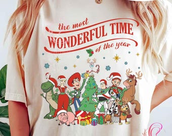 May include: A cream-colored t-shirt featuring a festive design with the text "the most WONDERFUL TIME of the year" in red. The graphic includes cartoon characters, a Christmas tree, and snowflakes, perfect for the holiday season.