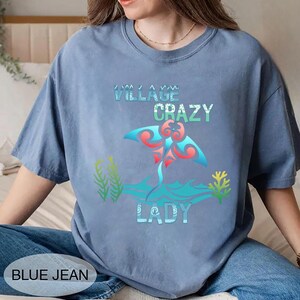 K&ouml;nnte beinhalten: Hellblaues T-Shirt mit dem Text "VILLAGE CRAZY LADY" und einer farbenfrohen Rochen-Grafik. Die Grafik zeigt rote, blaue und t&uuml;rkisfarbene Elemente. Die Worte "BLUE JEAN" sind unten links im Bild aufgedruckt.