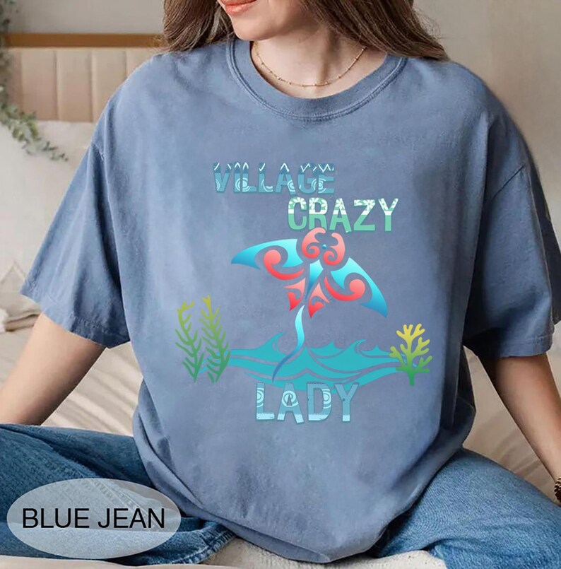 K&ouml;nnte beinhalten: Hellblaues T-Shirt mit dem Text "VILLAGE CRAZY LADY" und einer farbenfrohen Rochen-Grafik. Die Grafik zeigt rote, blaue und t&uuml;rkisfarbene Elemente. Die Worte "BLUE JEAN" sind unten links im Bild aufgedruckt.