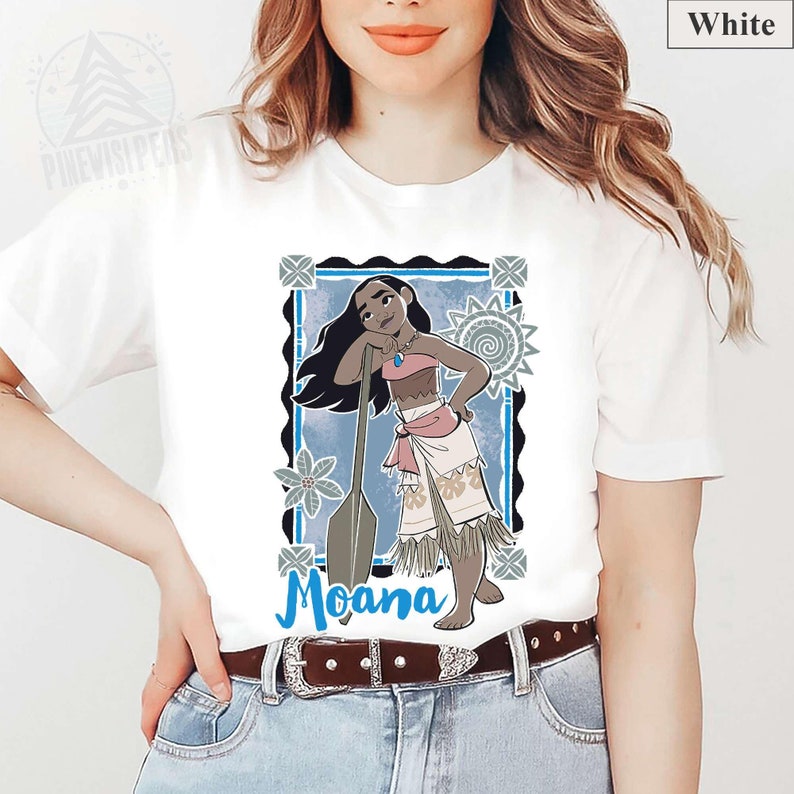 May include: White t-shirt with a graphic of Moana, framed in blue and black. The illustration depicts Moana with long black hair, a pink top, and a white skirt, leaning on a paddle. The word "Moana" is written in blue.