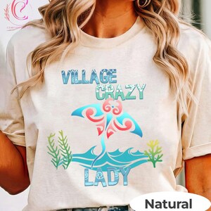 K&ouml;nnte beinhalten: Beiges T-Shirt mit den Worten "VILLAGE CRAZY LADY" in Blau und Gr&uuml;n, mit einem Mantarochen-Design in Rot und Blau. Das Shirt hat auch gr&uuml;ne und gelbe Seegras-Grafiken. Das Wort "Natural" steht unten.