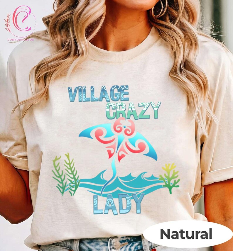 K&ouml;nnte beinhalten: Beiges T-Shirt mit den Worten "VILLAGE CRAZY LADY" in Blau und Gr&uuml;n, mit einem Mantarochen-Design in Rot und Blau. Das Shirt hat auch gr&uuml;ne und gelbe Seegras-Grafiken. Das Wort "Natural" steht unten.