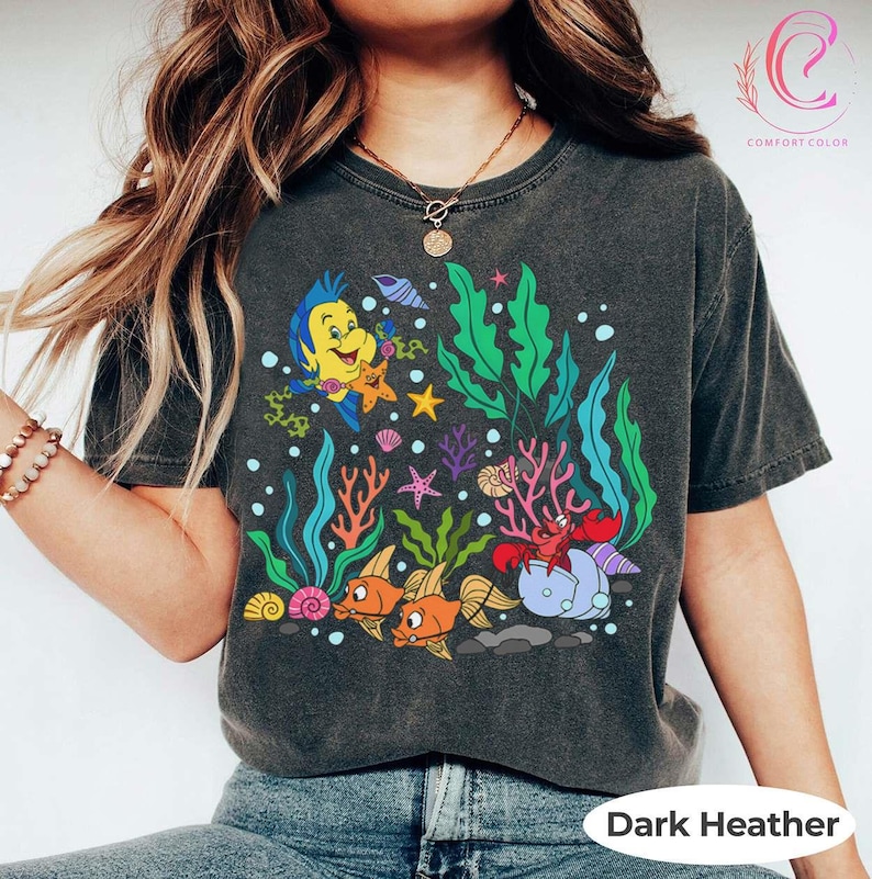 K&ouml;nnte beinhalten: Dunkelgraues T-Shirt mit einem farbenfrohen Unterwassermotiv mit Zeichentrickfischen, Korallen und einer Krabbe. Das Design enth&auml;lt den Text "Dark Heather" unten. Das Shirt ist von der Marke Comfort Colors.