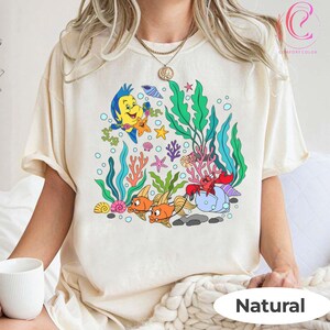 K&ouml;nnte beinhalten: Ein cremefarbenes T-Shirt mit einer farbenfrohen Cartoon-Unterwasserszene. Das Design zeigt gelbe Fische, eine Krabbe und andere Meeresbewohner, mit dem Wort "Natural" unten aufgedruckt. Das Shirt besteht aus Comfort Color-Material.