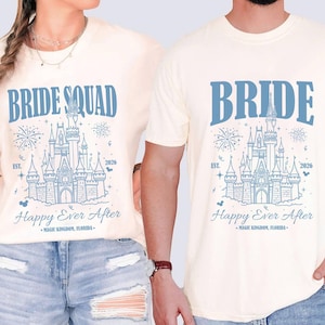 Può includere: Due magliette color crema con una grafica blu di un castello con fuochi d'artificio. La maglietta a sinistra riporta la scritta BRIDE SQUAD, quella a destra BRIDE. Entrambe includono il testo EST. 2026, Happy Ever After e MAGIC KINGDOM, FLORIDA.