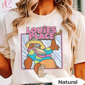 May include: A natural-coloured t-shirt with a cartoon monkey wearing a hat, scarf, and a blue shirt with polka dots. The text "Louie's Place" is above the monkey in pink and white letters. The word "Natural" is at the bottom.