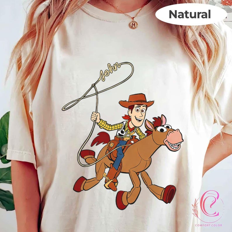 May include: Cream-colored t-shirt featuring a cartoon illustration of a cowboy riding a horse. The cowboy is wearing a brown hat, yellow shirt, and blue pants. The horse is brown with a red saddle and hooves. The word "John" is written above the cowboy.