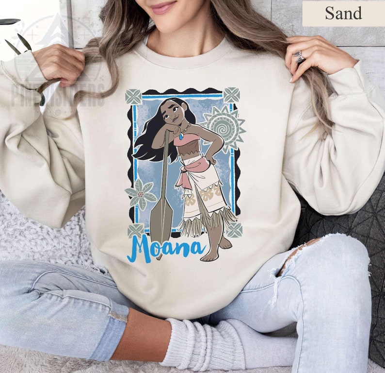 May include: Sand-coloured sweatshirt featuring a graphic of Moana, with a blue background and the word "Moana" in blue script. The design includes a cartoon illustration of Moana with a paddle. The sweatshirt is a neutral colour.