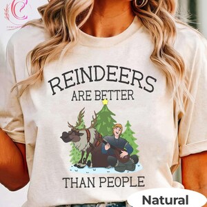 May include: A beige t-shirt with the text "REINDEERS ARE BETTER THAN PEOPLE" in black. The graphic features a cartoon reindeer, a Christmas tree, and a man playing a lute. The word "Natural" is printed at the bottom.