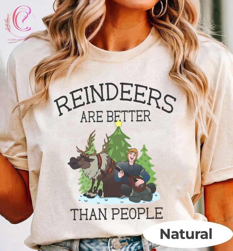 May include: A beige t-shirt with the text "REINDEERS ARE BETTER THAN PEOPLE" in black. The graphic features a cartoon reindeer, a Christmas tree, and a man playing a lute. The word "Natural" is printed at the bottom.