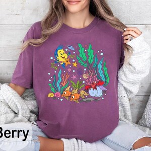 K&ouml;nnte beinhalten: Beerenfarbenes T-Shirt mit einer Cartoon-Grafik von Unterwasser-Meereslebewesen. Das Design umfasst Fische, eine Krabbe, Seetang und andere Meeresbewohner. Das Wort "Berry" ist unten in schwarzer Schrift gedruckt.