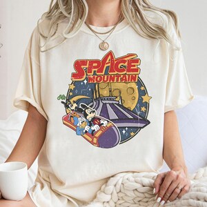 以下が含まれることがあります： クリーム色のTシャツに、ミッキーマウスとグーフィーがスペースマウンテンに乗っているヴィンテージスタイルのグラフィックがプリントされています。 「SPACE MOUNTAIN」の文字は赤と青の文字でプリントされています。