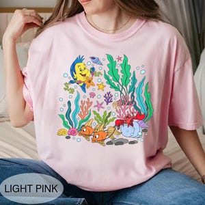 K&ouml;nnte beinhalten: Hellrosa T-Shirt mit einer farbenfrohen Cartoon-Grafik einer Unterwasserszene mit Fischen, Seetang und Meeresbewohnern. Der Text "LIGHT PINK" befindet sich am unteren Rand des Shirts.
