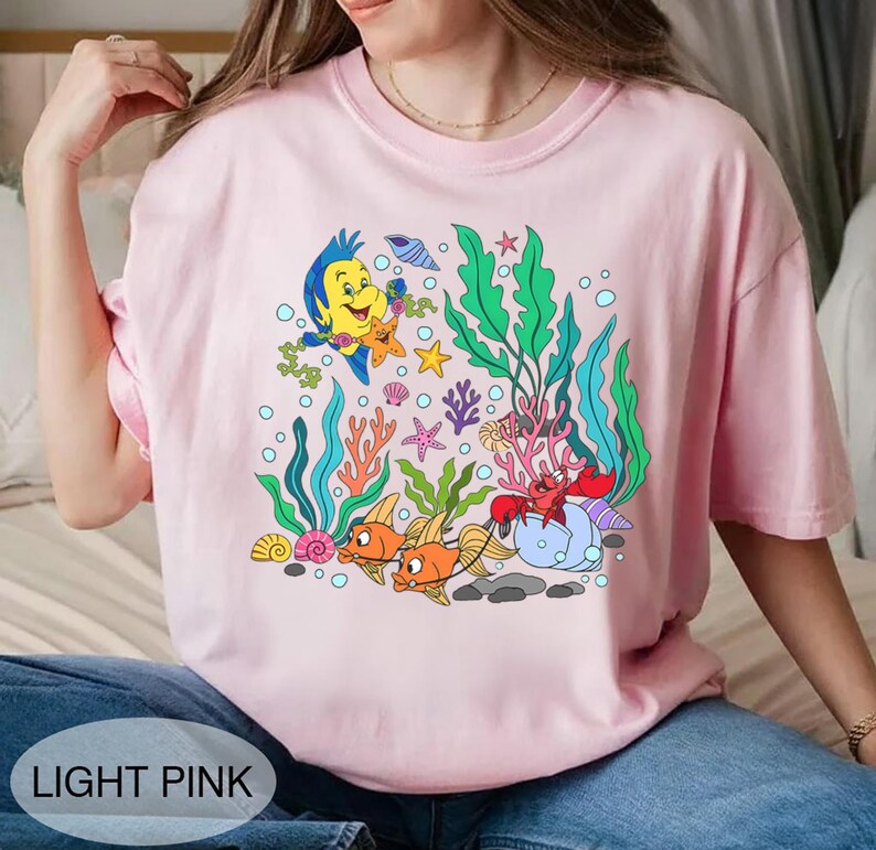 K&ouml;nnte beinhalten: Hellrosa T-Shirt mit einer farbenfrohen Cartoon-Grafik einer Unterwasserszene mit Fischen, Seetang und Meeresbewohnern. Der Text "LIGHT PINK" befindet sich am unteren Rand des Shirts.