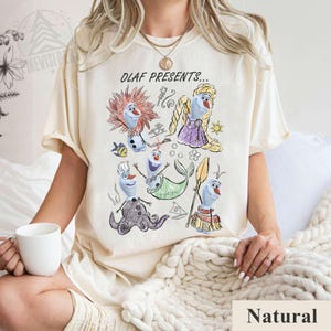 Puede incluir: Camiseta de color crema con un gr&aacute;fico colorido de Olaf de Frozen con varios disfraces, con el texto "OLAF PRESENTS...". La camiseta est&aacute; hecha de un material suave y tiene mangas cortas.