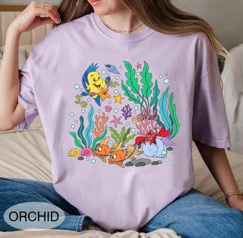 K&ouml;nnte beinhalten: Helllila T-Shirt mit einem farbenfrohen Cartoon-Motiv von Meeresbewohnern, darunter Fische, eine Krabbe und Seetang. Das Wort "ORCHID" ist unten aufgedruckt. Das T-Shirt ist aus weichem Material.