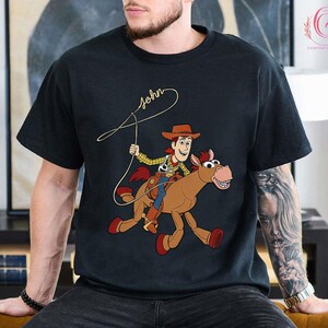 May include: Black t-shirt featuring a cartoon illustration of a cowboy riding a horse. The cowboy is wearing a brown hat, yellow shirt, and blue pants. The horse is brown with a red saddle. The word "John" is written in cursive above the cowboy.