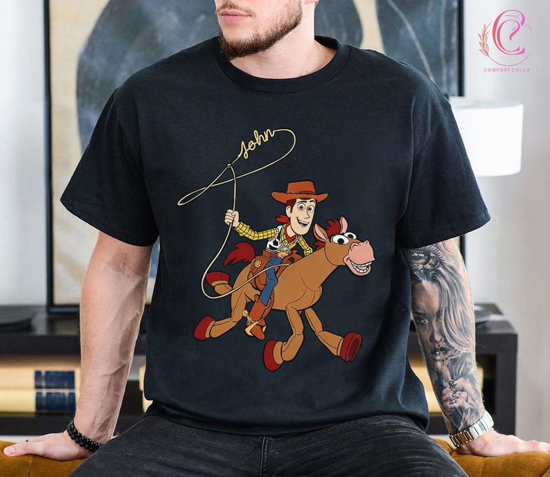 May include: Black t-shirt featuring a cartoon illustration of a cowboy riding a horse. The cowboy is wearing a brown hat, yellow shirt, and blue pants. The horse is brown with a red saddle. The word "John" is written in cursive above the cowboy.