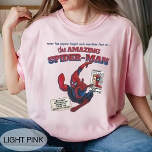 Op de afbeelding: Lichtroze T-shirt met een stripboekachtige afbeelding van Spider-Man in actie. De tekst "the AMAZING SPIDER-MAN" staat boven de illustratie. Extra tekst bevat "never has anyone fought such merciless foes as..." en "ANOTHER SPECTACULAR SMASH-HIT FROM THE HOUSE OF IDEAS!"