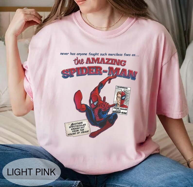 Op de afbeelding: Lichtroze T-shirt met een stripboekachtige afbeelding van Spider-Man in actie. De tekst "the AMAZING SPIDER-MAN" staat boven de illustratie. Extra tekst bevat "never has anyone fought such merciless foes as..." en "ANOTHER SPECTACULAR SMASH-HIT FROM THE HOUSE OF IDEAS!"