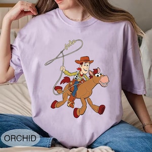 May include: Orchid-colored t-shirt featuring a cartoon illustration of a cowboy riding a horse, with the name "John" written above. The cowboy wears a brown hat, yellow shirt, and blue pants. The horse is brown with red hooves.
