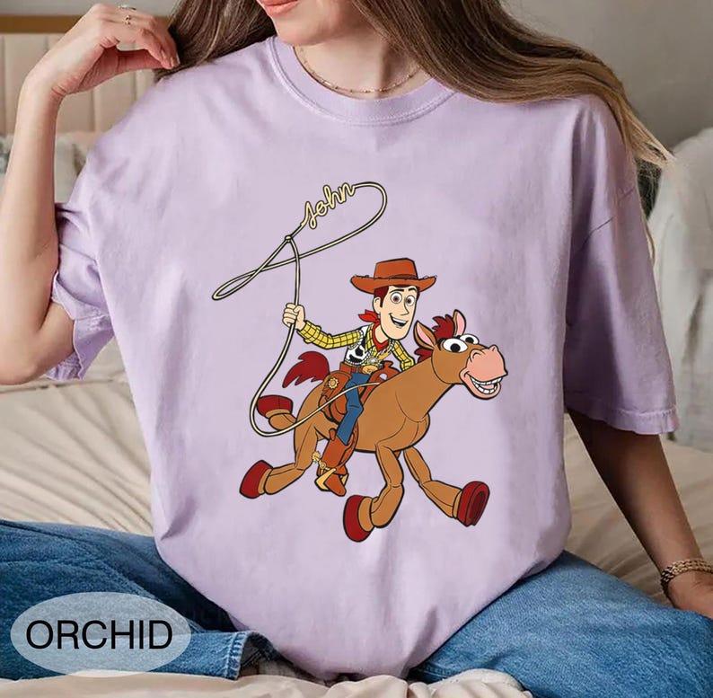 May include: Orchid-colored t-shirt featuring a cartoon illustration of a cowboy riding a horse, with the name "John" written above. The cowboy wears a brown hat, yellow shirt, and blue pants. The horse is brown with red hooves.