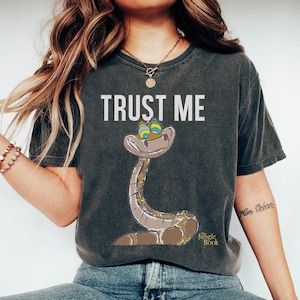 May include: A dark gray t-shirt with the text "TRUST ME" and a cartoon image of Kaa the snake from Disney's The Jungle Book. The snake has green eyes and a sly smile.