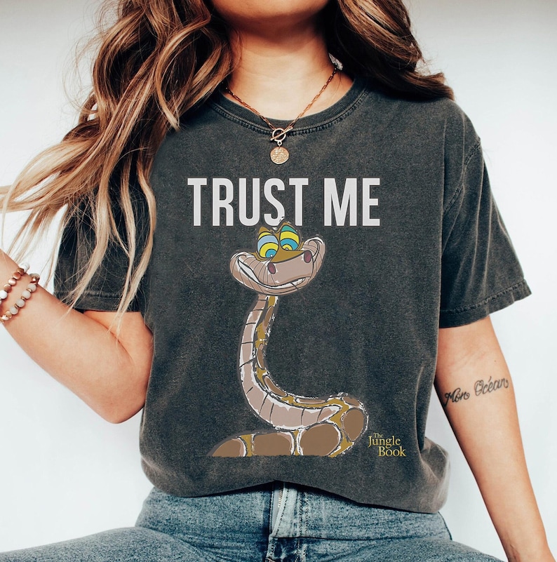 May include: A dark gray t-shirt with the text "TRUST ME" and a cartoon image of Kaa the snake from Disney's The Jungle Book. The snake has green eyes and a sly smile.