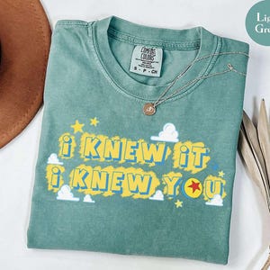 May include: A light green short-sleeve t-shirt featuring the text I KNEW IT I KNEW YOU in yellow and blue lettering. The design includes small white clouds, yellow stars, and a red star icon. The shirt has a Comfort Colors brand label.