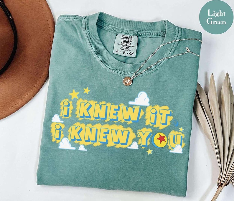 May include: A light green short-sleeve t-shirt featuring the text I KNEW IT I KNEW YOU in yellow and blue lettering. The design includes small white clouds, yellow stars, and a red star icon. The shirt has a Comfort Colors brand label.